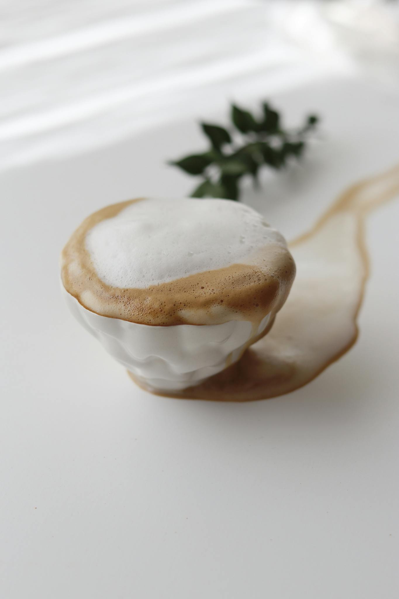 A coffee cup overflowing with foam, captured from above on a white surface with a sprig decoration.