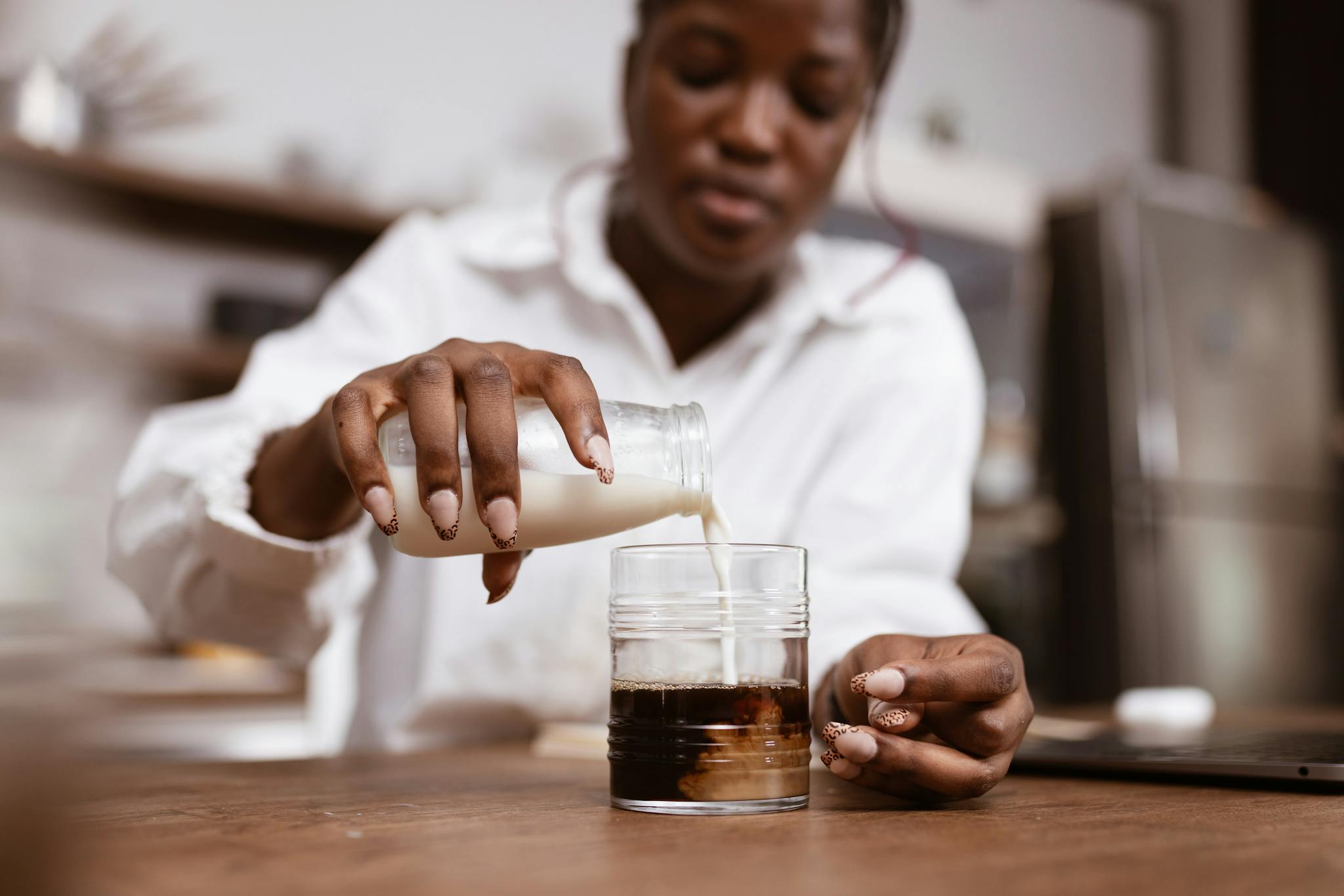 A woman pours milk into a glass of coffee on a wooden table, focused on brewing.