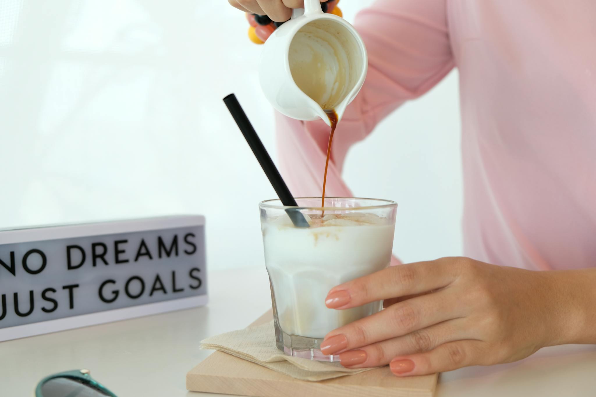 Close-up of a hand pouring coffee into a glass of milk with a motivational sign in the background.